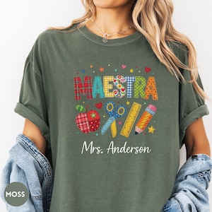 May include: A moss green t-shirt featuring the word MAESTRA in colorful patterned block letters, accompanied by graphics of a plaid apple, blue scissors, a yellow ruler, and a pencil, with the text Mrs. Anderson printed in white below.