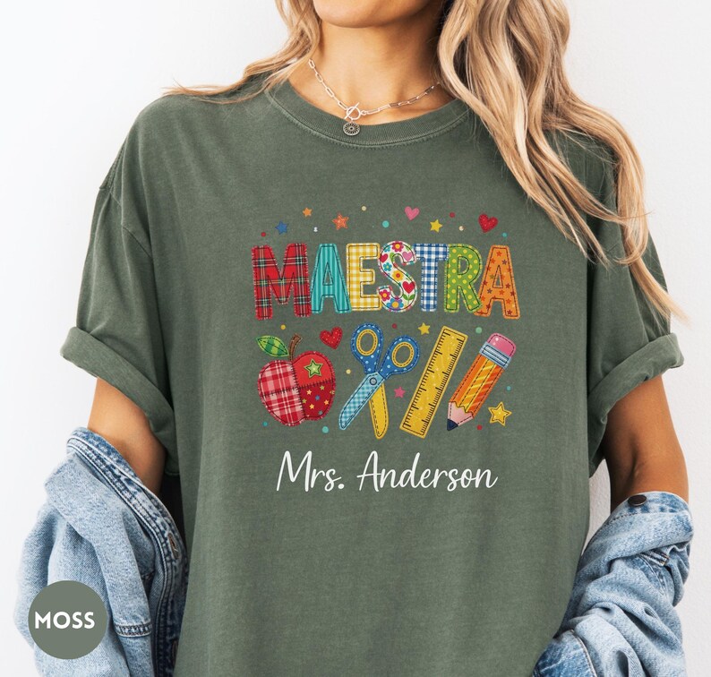 May include: A moss green t-shirt featuring the word MAESTRA in colorful patterned block letters, accompanied by graphics of a plaid apple, blue scissors, a yellow ruler, and a pencil, with the text Mrs. Anderson printed in white below.