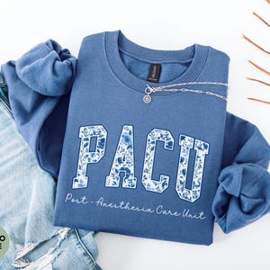 May include: Indigo blue sweatshirt with the letters "PACU" in a floral design. The text "Post - Anesthesia Care Unit" is below the letters. A silver necklace is draped across the sweatshirt, which is next to a pair of blue jeans.