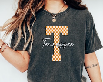 May include: Dark gray t-shirt featuring a large orange and white checkered "T" design, with the word "Tennessee" in cursive script below. The shirt is a comfortable, casual style.