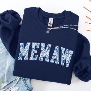 May include: Navy blue sweatshirt with the word "MEMAW" in white letters filled with a blue floral pattern. A silver necklace with a pendant is draped across the sweatshirt. The sweatshirt is folded next to a pair of blue jeans.
