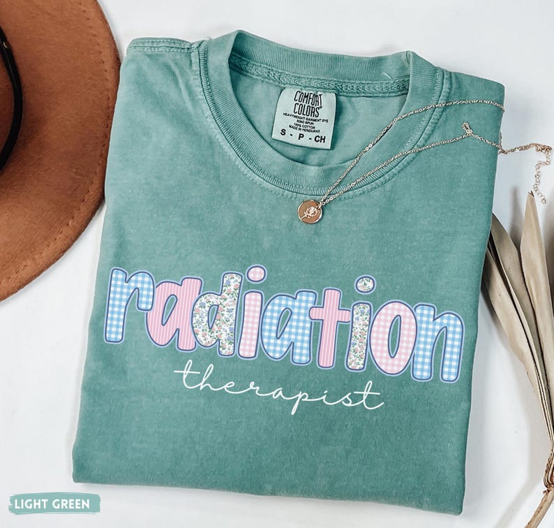 May include: A light green t-shirt with the words "radiation therapist" in a decorative font. The word "radiation" is in pink and blue gingham and floral patterns. A silver necklace with a small pendant rests on the shirt.