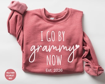 May include: A heather scarlet red crewneck sweatshirt featuring white text that reads I GO BY grammy NOW with a small heart icon and the text Est. 2026 below. The garment has long sleeves with ribbed cuffs and a round neckline.