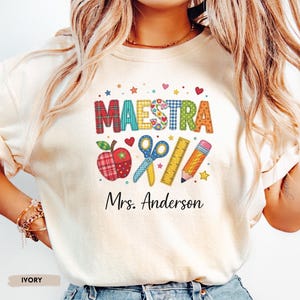 Personalized MAESTRA shirt with colorful school-themed letters, apple, scissors, ruler, pencil, and custom teacher name on an ivory Comfort Colors tee.