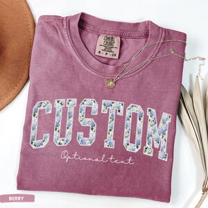 May include: Berry-colored t-shirt with the word "CUSTOM" in a floral design. The shirt has a crew neck and short sleeves. A delicate necklace with a pendant rests on the shirt. The words "Optional text" are below the word "CUSTOM".