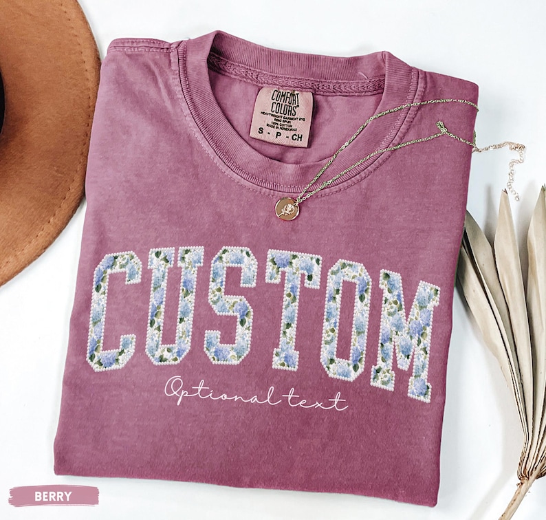 May include: Berry-colored t-shirt with the word "CUSTOM" in a floral design. The shirt has a crew neck and short sleeves. A delicate necklace with a pendant rests on the shirt. The words "Optional text" are below the word "CUSTOM".