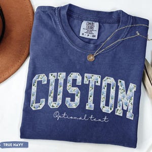 May include: A navy blue t-shirt with the word "CUSTOM" in white block letters filled with a blue floral pattern. The text "Optional text" is below the word "CUSTOM". A gold necklace with a pendant rests on the shirt.
