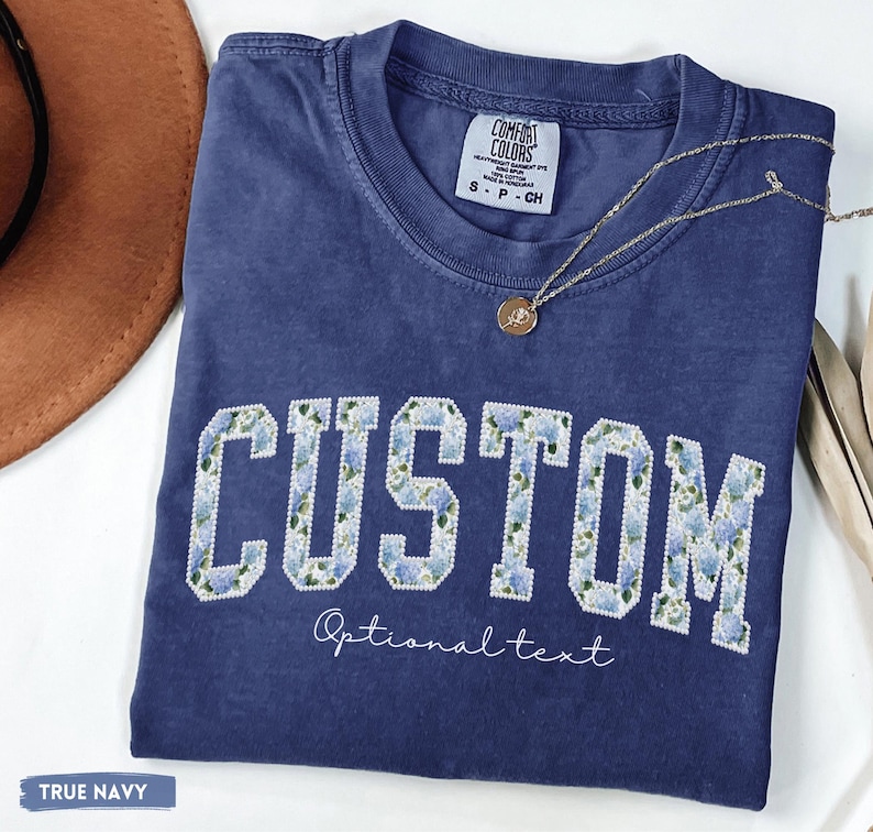 May include: A navy blue t-shirt with the word "CUSTOM" in white block letters filled with a blue floral pattern. The text "Optional text" is below the word "CUSTOM". A gold necklace with a pendant rests on the shirt.