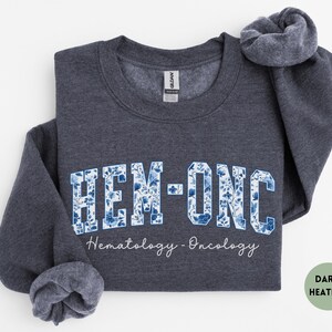May include: Dark heather gray sweatshirt with the text "HEM-ONC" in a blue floral design. The words "Hematology - Oncology" are written below the main text in a script font. The sweatshirt is folded.
