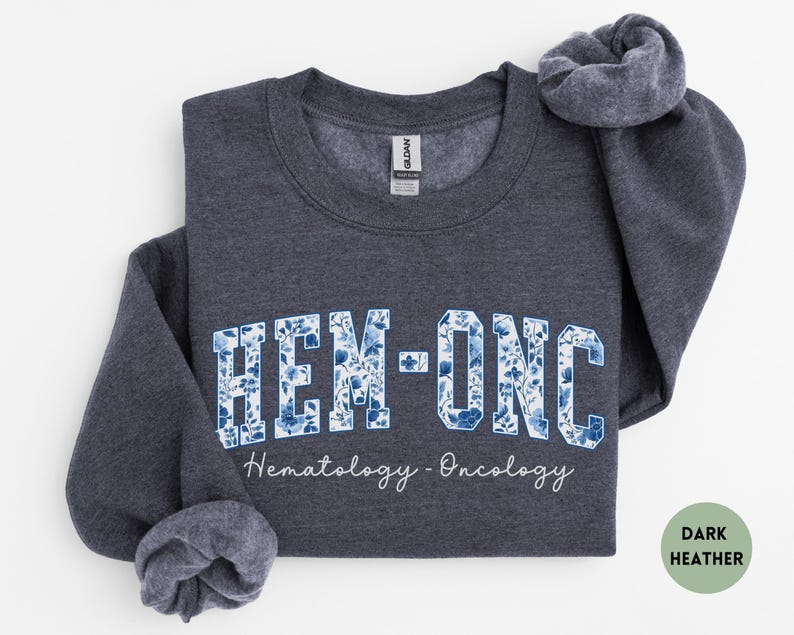 May include: Dark heather gray sweatshirt with the text "HEM-ONC" in a blue floral design. The words "Hematology - Oncology" are written below the main text in a script font. The sweatshirt is folded.