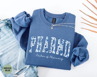 May include: An indigo blue crewneck sweatshirt featuring the text PHARMD in a collegiate font filled with a blue and white floral pattern, with the words Doctor of Pharmacy written below in a white script font.