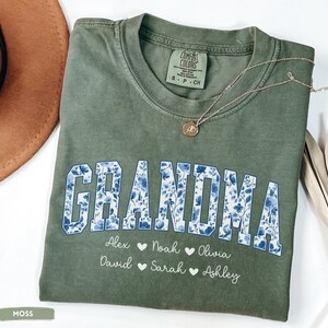 May include: A moss green t-shirt with the word "GRANDMA" in blue floral print. Below the word are names with hearts. A gold necklace with a pendant is on the shirt. The shirt is folded.