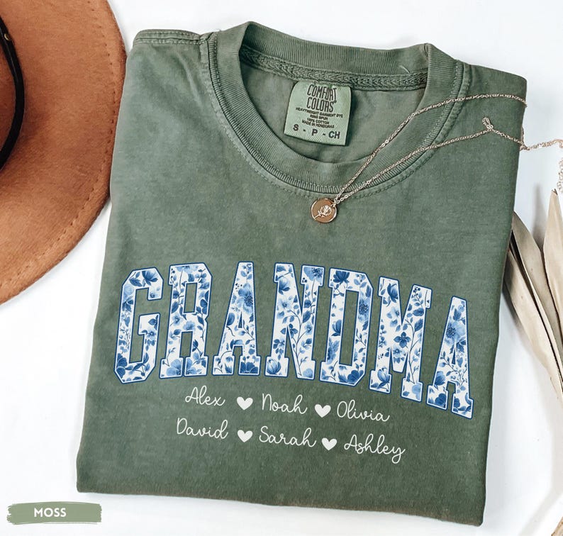 May include: A moss green t-shirt with the word "GRANDMA" in blue floral print. Below the word are names with hearts. A gold necklace with a pendant is on the shirt. The shirt is folded.