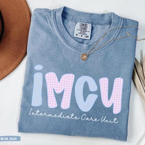 May include: Blue jean colored t-shirt with the letters "IMCU" in a combination of pink gingham and blue striped patterns. The words "Intermediate Care Unit" are printed below the letters. A gold necklace with a pendant is draped across the shirt.