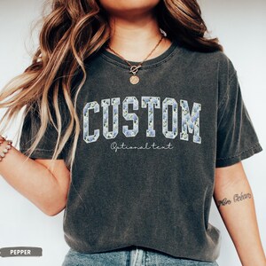 May include: A dark gray t-shirt with the word "CUSTOM" in a collegiate font, filled with a blue floral pattern. Below the word "CUSTOM" is the text "Optional text" in a script font. The shirt is a relaxed fit.