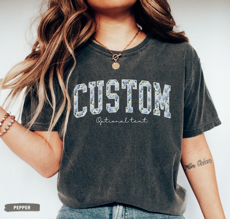 May include: A dark gray t-shirt with the word "CUSTOM" in a collegiate font, filled with a blue floral pattern. Below the word "CUSTOM" is the text "Optional text" in a script font. The shirt is a relaxed fit.