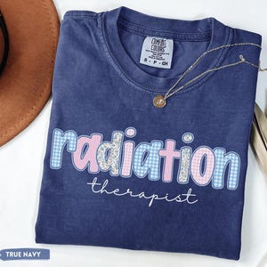 May include: A navy blue t-shirt with the words "radiation therapist" in a decorative font. The word "radiation" is in a pink and blue gingham pattern. The shirt has a Comfort Colors tag.