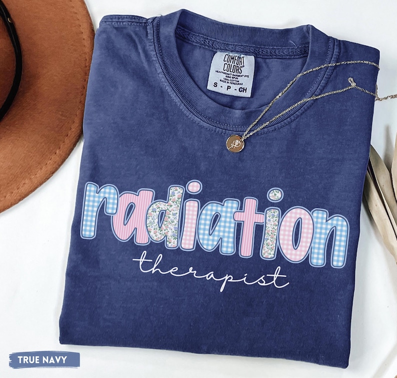 May include: A navy blue t-shirt with the words "radiation therapist" in a decorative font. The word "radiation" is in a pink and blue gingham pattern. The shirt has a Comfort Colors tag.