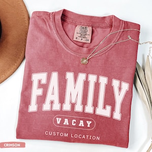 Peut inclure: Un t-shirt &agrave; manches courtes couleur cramoisi avec un col rond. Le devant pr&eacute;sente le mot FAMILY en grandes lettres blanches, VACAY dans un ovale et le texte CUSTOM LOCATION en dessous. Un petit collier dor&eacute; repose sur le col.