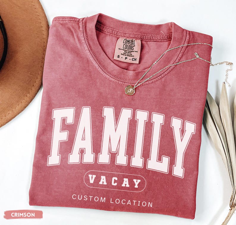 Peut inclure: Un t-shirt &agrave; manches courtes couleur cramoisi avec un col rond. Le devant pr&eacute;sente le mot FAMILY en grandes lettres blanches, VACAY dans un ovale et le texte CUSTOM LOCATION en dessous. Un petit collier dor&eacute; repose sur le col.
