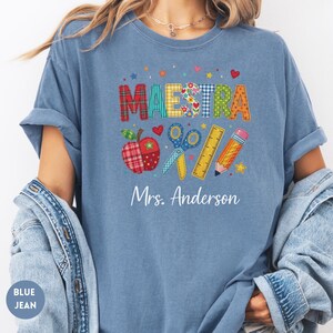 May include: A blue t-shirt featuring the word MAESTRA in colorful patterned letters above illustrations of an apple, scissors, a ruler, and a pencil. Below these graphics, the text Mrs. Anderson is printed in white script on the chest.