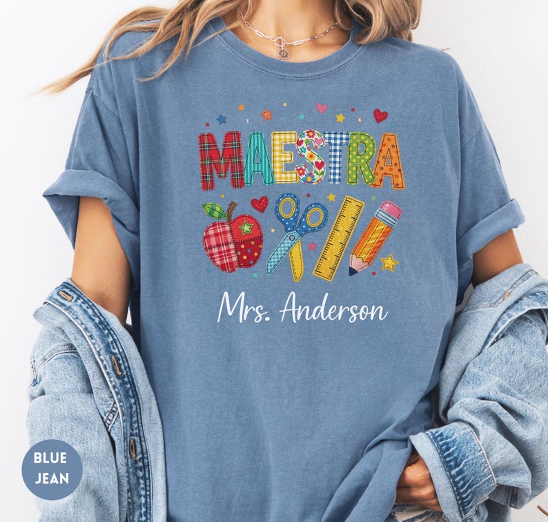 May include: A blue t-shirt featuring the word MAESTRA in colorful patterned letters above illustrations of an apple, scissors, a ruler, and a pencil. Below these graphics, the text Mrs. Anderson is printed in white script on the chest.