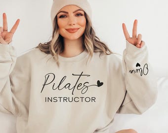 May include: A beige crewneck sweatshirt featuring the black text Pilates INSTRUCTOR with a small heart icon on the front, and the name Olivia with a heart icon printed in black on the left sleeve cuff, made of a soft, solid-colored fabric.