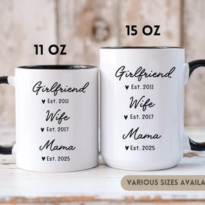 May include: Two white ceramic coffee mugs with black handles and black rims. The mugs have black text that reads "Girlfriend Est. 2011 Wife Est. 2017 Mama Est. 2025". The mugs are available in 11 oz and 15 oz sizes.