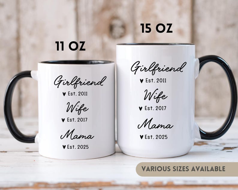 May include: Two white ceramic coffee mugs with black handles and black rims. The mugs have black text that reads "Girlfriend Est. 2011 Wife Est. 2017 Mama Est. 2025". The mugs are available in 11 oz and 15 oz sizes.