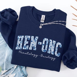 May include: Navy blue sweatshirt with the text "HEM-ONC" in a floral design. The words "Hematology - Oncology" are written below the main text in a script font. A silver necklace with a circular pendant is draped across the sweatshirt.