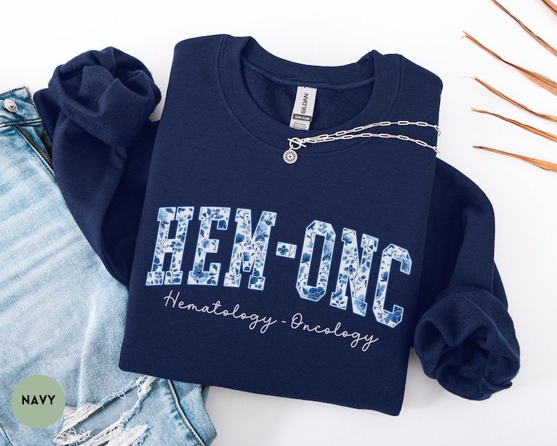 May include: Navy blue sweatshirt with the text "HEM-ONC" in a floral design. The words "Hematology - Oncology" are written below the main text in a script font. A silver necklace with a circular pendant is draped across the sweatshirt.