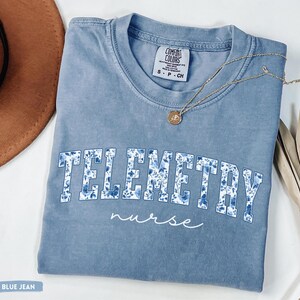 May include: A blue jean Comfort Colors t-shirt with the word "TELEMETRY" in large, floral-patterned letters and the word "nurse" in script. A gold necklace with a pendant rests on the shirt.