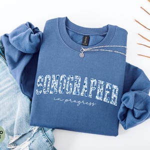 May include: A blue sweatshirt with the word "SONOGRAPHER" in a floral design and the words "in progress" in a script font. A silver necklace is draped across the sweatshirt. The sweatshirt is paired with blue jeans.
