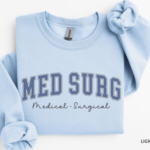 May include: Light blue sweatshirt with the text "MED SURG" in a navy blue collegiate font, with "Medical - Surgical" written below in a script font. The sweatshirt is folded, with the sleeves rolled up.