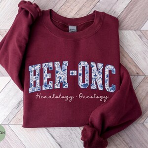 May include: Maroon sweatshirt with the text "HEM-ONC" in white letters filled with a blue floral pattern. Below the text is the script "Hematology - Oncology". The word "MAROON" is in a green circle in the lower left corner.