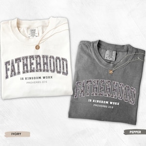 May include: Two t-shirts, one ivory and one pepper, with the word "FATHERHOOD" in a collegiate font. The text "IS KINGDOM WORK PROVERBS 22:6" is below. Each shirt has a gold pendant necklace. The ivory shirt is on the left, and the pepper shirt is on the right.