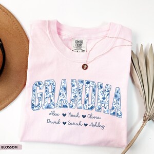 May include: A pale pink t-shirt featuring the word "GRANDMA" in a blue floral design. Underneath are names: Alex, Noah, Olivia, David, Sarah, and Ashley. A gold necklace with a pendant is placed on the shirt.