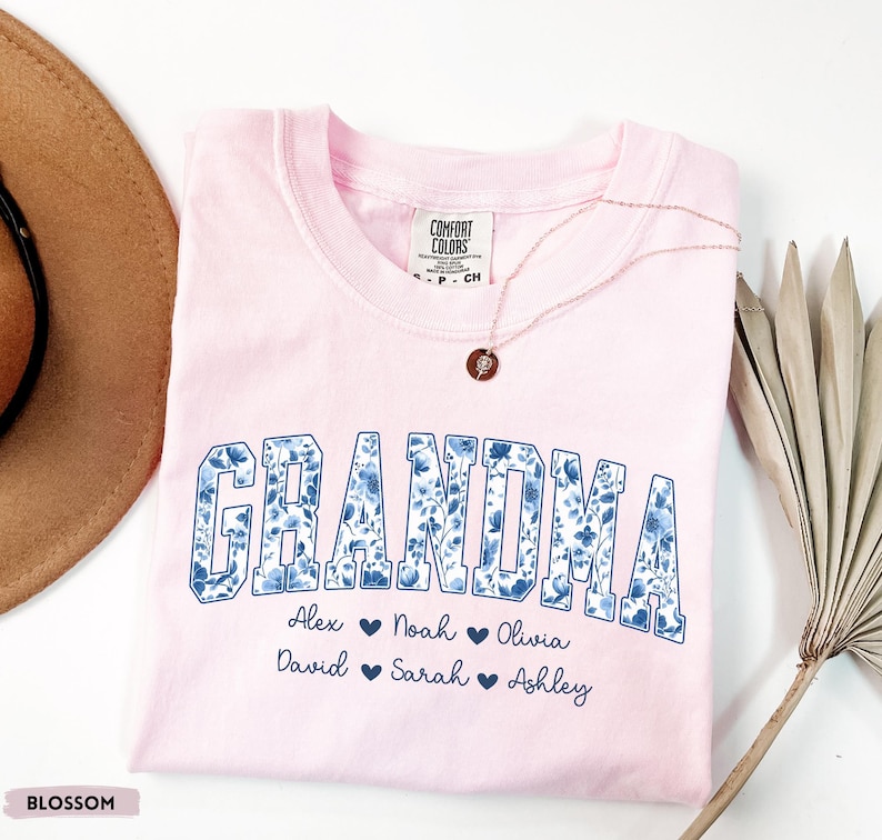 May include: A pale pink t-shirt featuring the word "GRANDMA" in a blue floral design. Underneath are names: Alex, Noah, Olivia, David, Sarah, and Ashley. A gold necklace with a pendant is placed on the shirt.