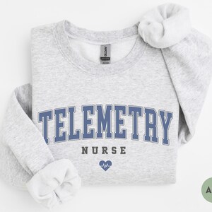 May include: Ash-colored sweatshirt with the word "TELEMETRY" in blue block letters, with "NURSE" below and a heart with a heartbeat symbol. The sweatshirt has a crew neck and long sleeves.