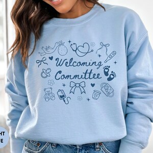 May include: Light blue sweatshirt with the words "Welcoming Committee" and baby-themed illustrations. The design includes a stork, baby feet, a stethoscope, and a baby bottle. The sweatshirt is a solid light blue color.