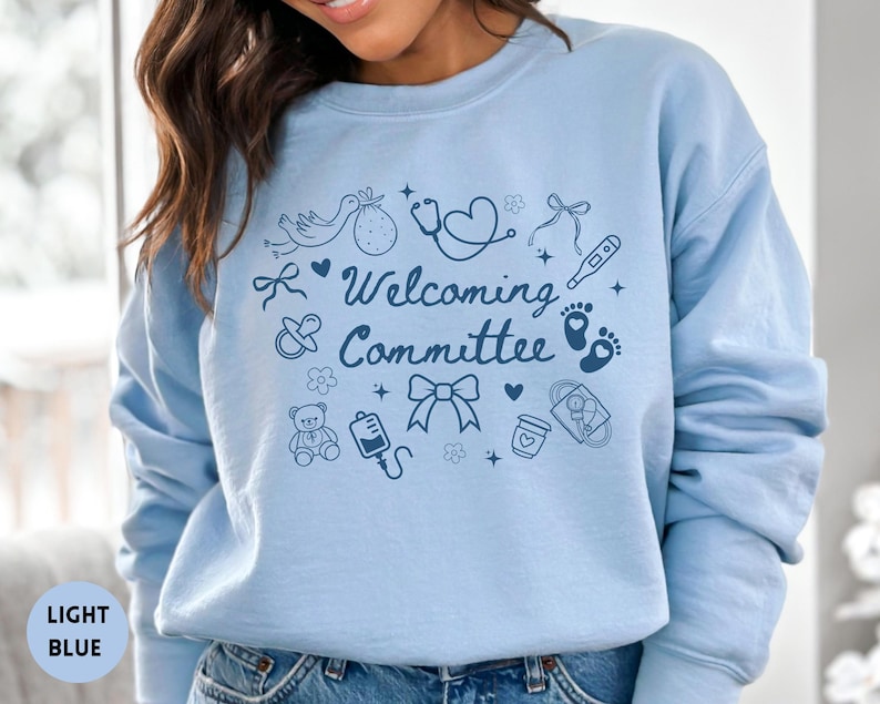 May include: Light blue sweatshirt with the words "Welcoming Committee" and baby-themed illustrations. The design includes a stork, baby feet, a stethoscope, and a baby bottle. The sweatshirt is a solid light blue color.