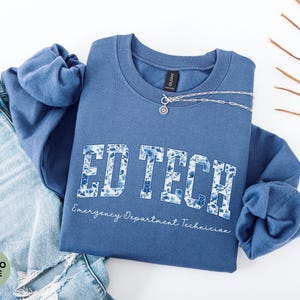 May include: Indigo blue crewneck sweatshirt with the text "ED TECH" in a floral design. The words "Emergency Department Technician" are written below the main text. A silver necklace is draped across the sweatshirt, which is paired with blue jeans.