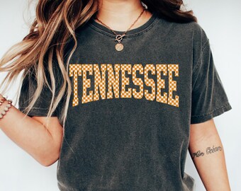 May include: A charcoal grey short-sleeve t-shirt featuring the word TENNESSEE printed in a bold, arched, orange and white checkered pattern across the chest. The shirt has a crew neckline and a slightly faded, vintage-inspired fabric texture.