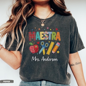 May include: A charcoal gray short-sleeve t-shirt featuring the word MAESTRA in colorful patterned letters. Below the text are graphics of a plaid apple, blue scissors, a yellow ruler, and a pencil. The name Mrs. Anderson is printed in white.