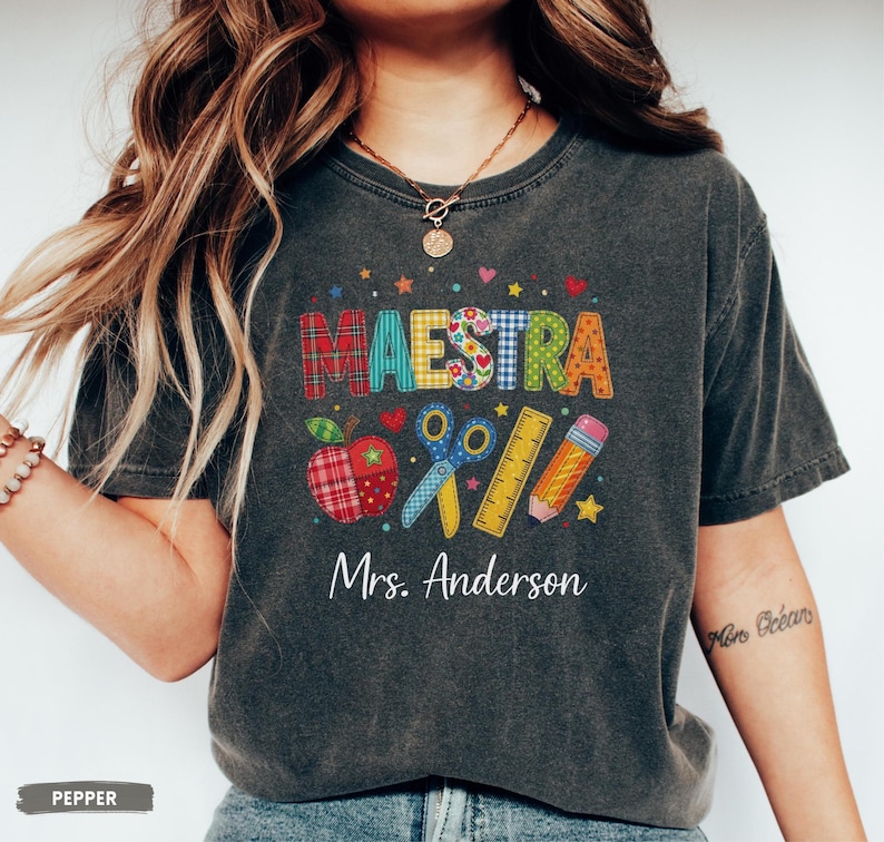 May include: A charcoal gray short-sleeve t-shirt featuring the word MAESTRA in colorful patterned letters. Below the text are graphics of a plaid apple, blue scissors, a yellow ruler, and a pencil. The name Mrs. Anderson is printed in white.