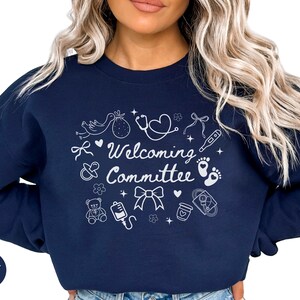 May include: Navy blue sweatshirt with the words "Welcoming Committee" in white script, surrounded by baby-themed illustrations. The design includes a stork, baby feet, a teddy bear, and a stethoscope.
