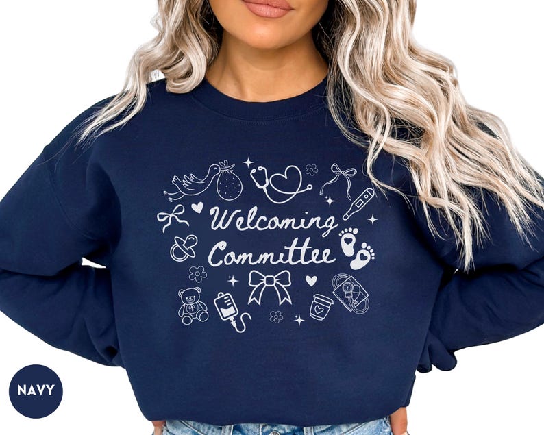 May include: Navy blue sweatshirt with the words "Welcoming Committee" in white script, surrounded by baby-themed illustrations. The design includes a stork, baby feet, a teddy bear, and a stethoscope.