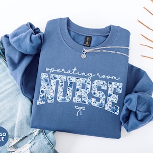May include: Indigo blue sweatshirt with the words "operating room NURSE" in white script and floral print. A silver necklace and a pair of blue jeans are next to the sweatshirt. The sweatshirt has long sleeves and a crew neck.