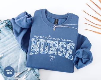 May include: Indigo blue sweatshirt with the words "operating room NURSE" in white script and floral print. A silver necklace and a pair of blue jeans are next to the sweatshirt. The sweatshirt has long sleeves and a crew neck.
