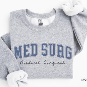 May include: Sport grey sweatshirt with the text "MED SURG" in blue block letters with a white outline. Below the text is "Medical - Surgical" in a cursive font. The sweatshirt has white fleece lining the collar and cuffs.